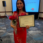 Jessica On stands in front of a screen with roses and a certificate.